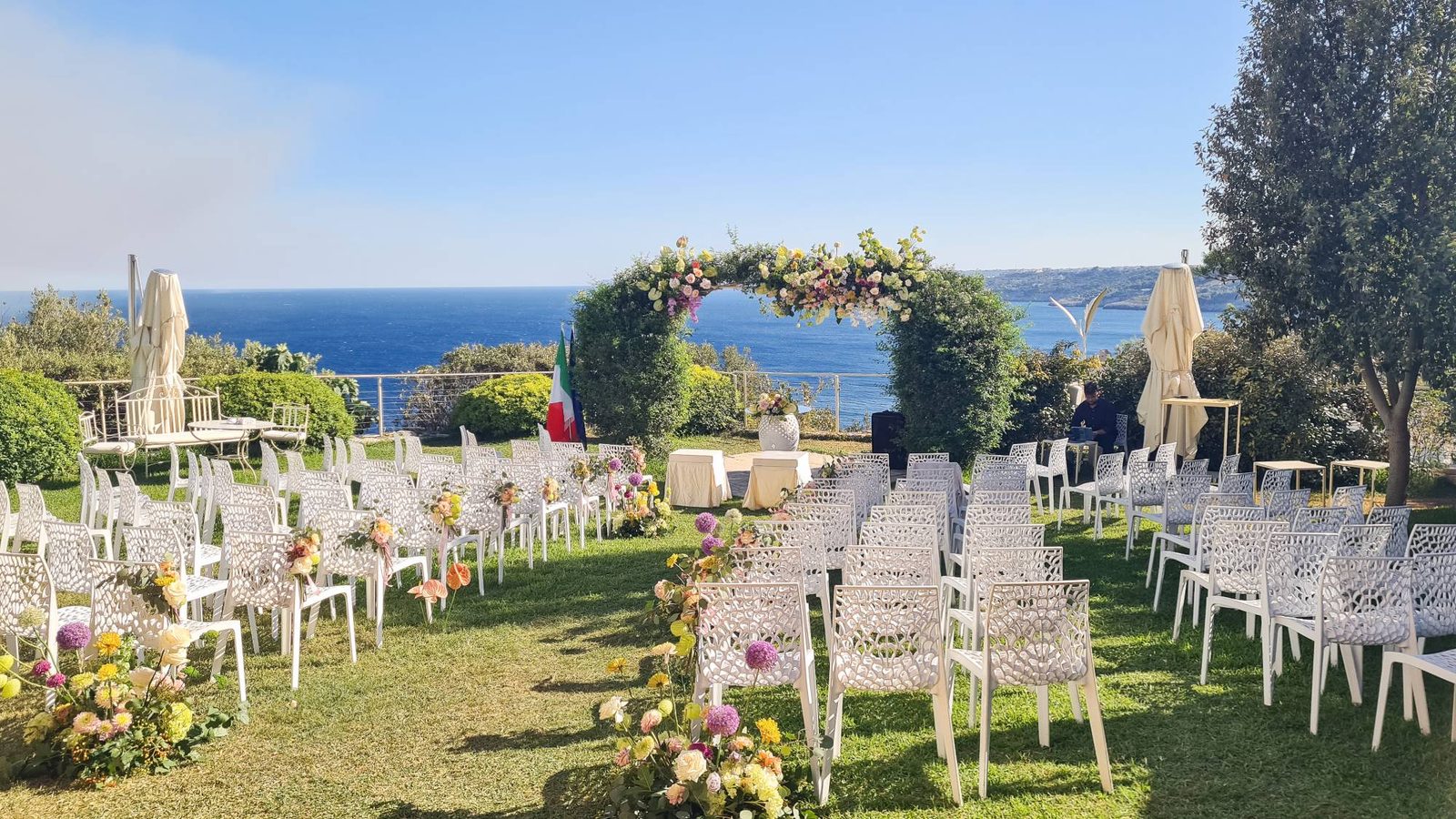 Allestimento per la cerimonia sul prato affacciato sul mare, con arco floreale colorato e sedie bianche — Cala dei Balcani, Salento