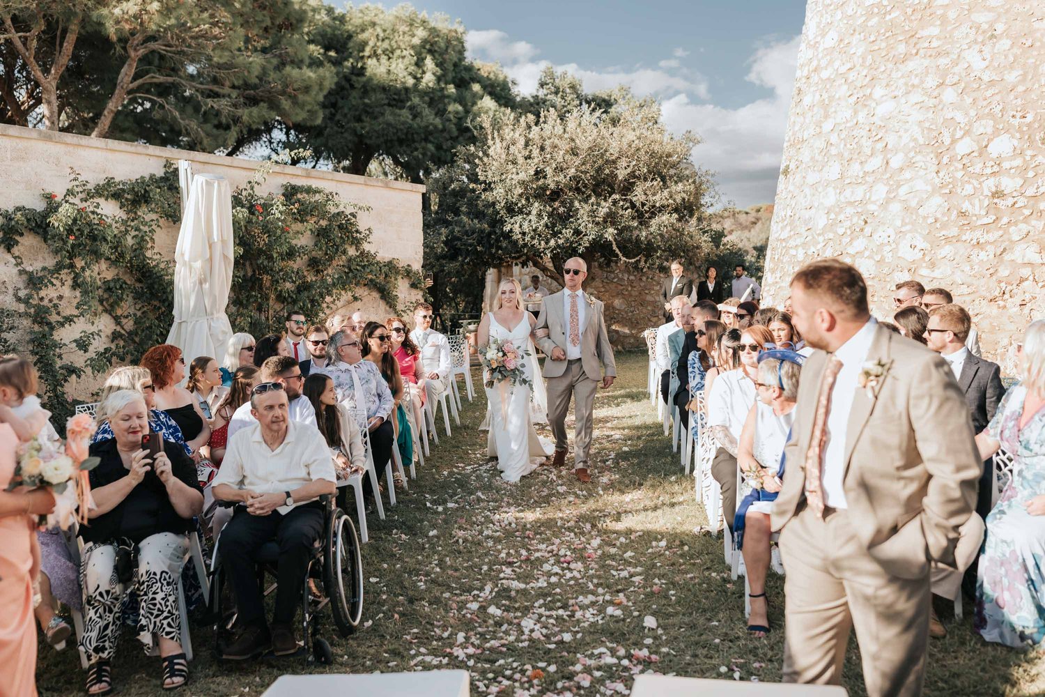 Invitados internacionales a lo largo del pasillo y el novio esperando a la novia, con la torre sarracena al fondo — Cala dei Balcani, Salento