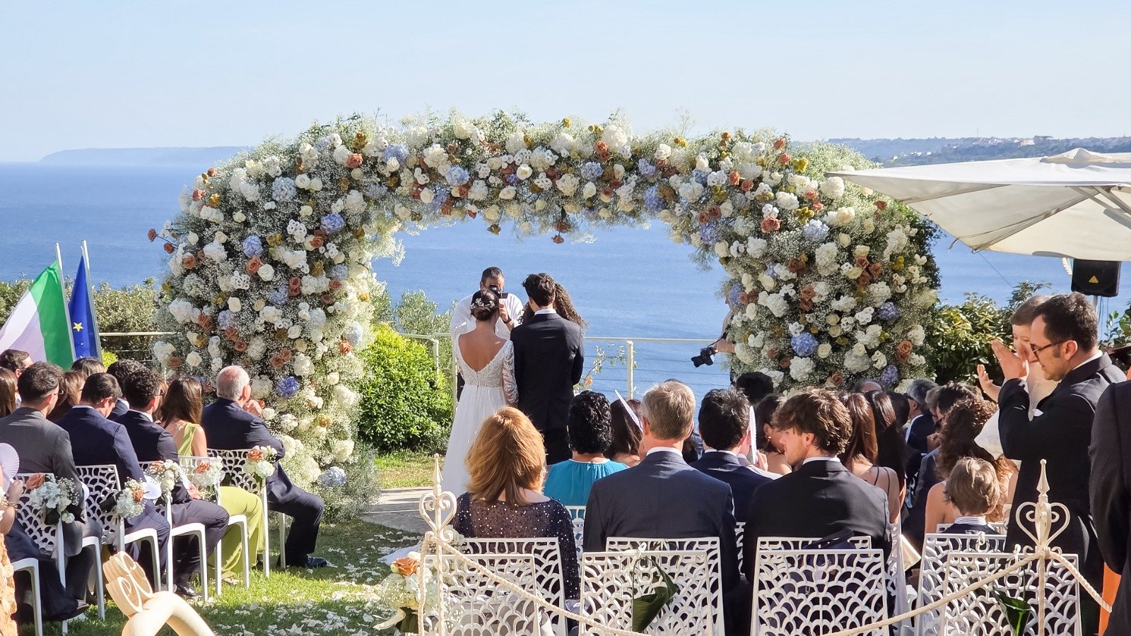 Los novios bajo el arco floral durante la ceremonia al aire libre con vistas al mar — Cala dei Balcani, Santa Cesarea Terme, Salento