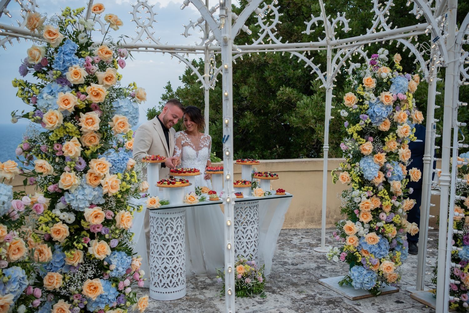 Sposi al buffet dei dolci sotto il gazebo decorato con rose pesca e ortensie azzurre, affacciato sul mare — Cala dei Balcani, Salento