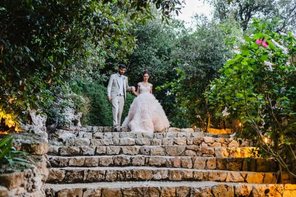 Gli sposi scendono la scalinata in pietra del giardino mediterraneo, tra lecci, pini e piante della macchia — Cala dei Balcani, Santa Cesarea Terme, Salento