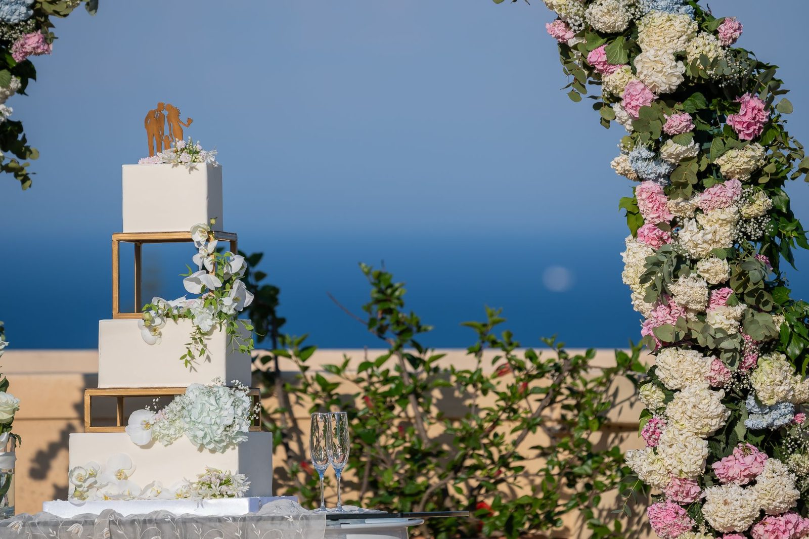 Torta nuziale a piani bianca e oro incorniciata da un arco di ortensie rosa e bianche, con il mare Adriatico sullo sfondo — Cala dei Balcani, Salento